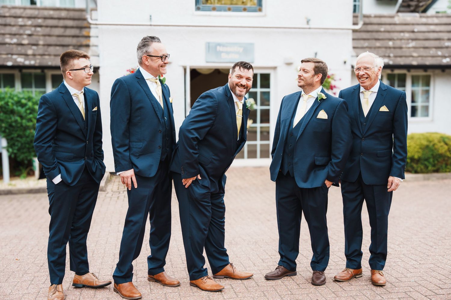 Groomsmen in navy suits and brown shoes pose casually outside a building.