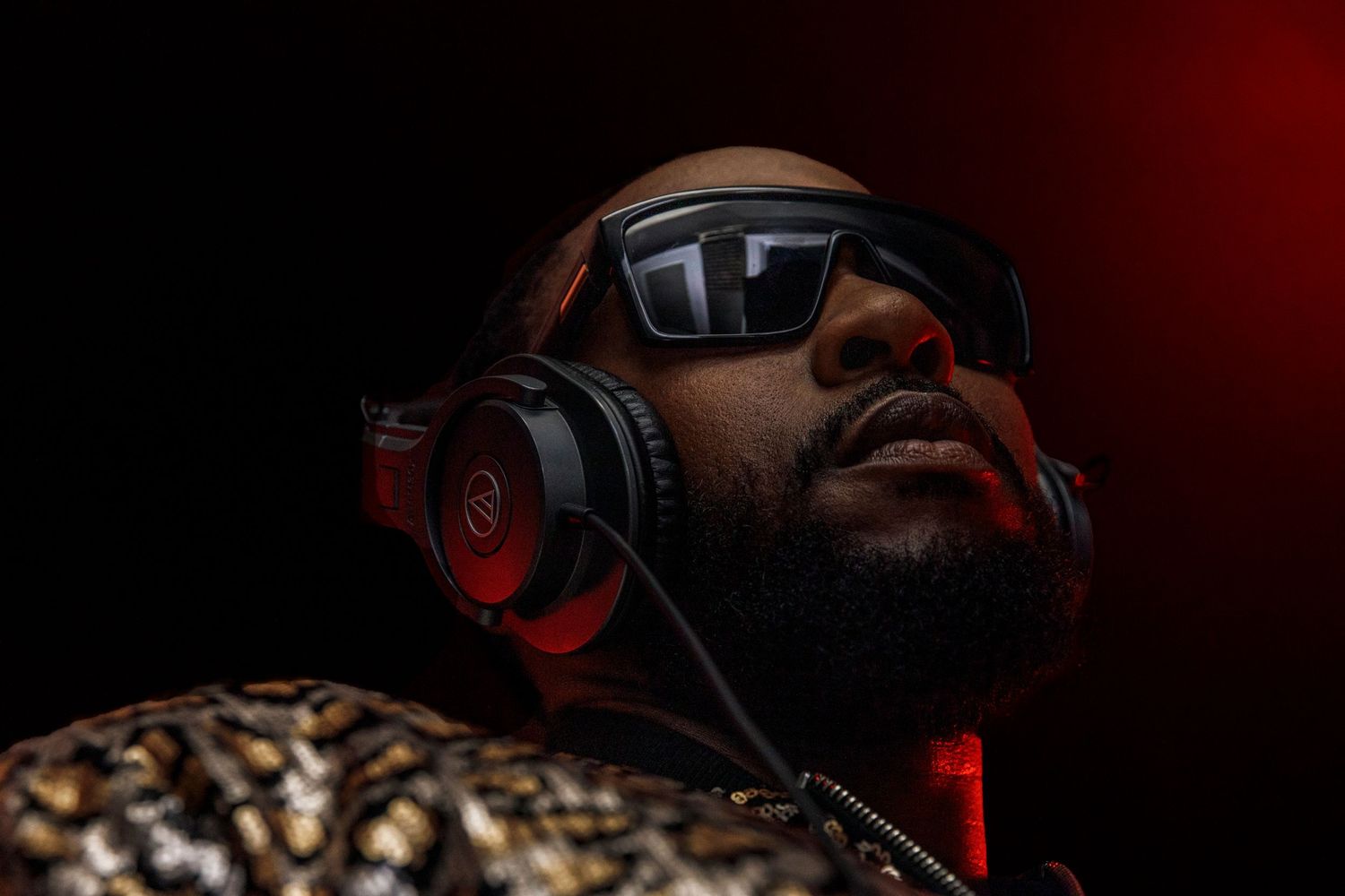 DJ wearing sunglasses and headphones illuminated by dramatic red lighting during a performance.