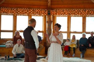 String lights decorate the wooden beams of a rustic event space filled with guests.