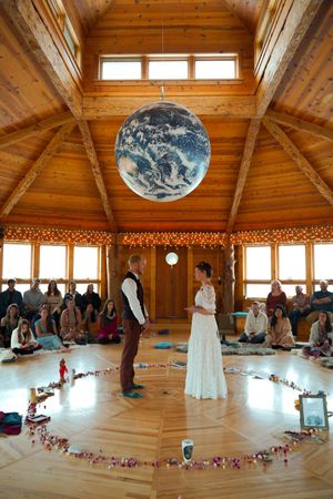 A wedding ceremony in a wooden octagonal room with a large Earth globe hanging from the ceiling and floral decorations.