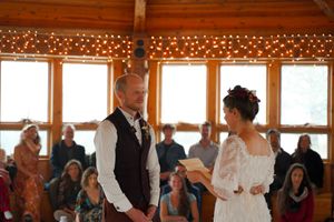 Wedding ceremony taking place in a rustic wooden venue with string lights and large windows.