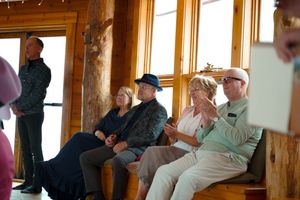 Wedding guests seated on benches by large windows in a rustic wooden venue.
