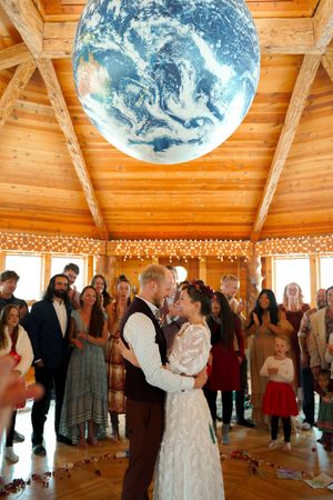Couple shares their first dance beneath a large illuminated Earth globe in a rustic wooden pavilion with string lights.