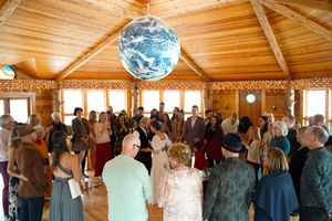 Wide angle shot of a wooden event hall with guests gathered for a wedding ceremony under string lights.