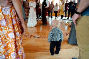Young child in plaid shirt watches wedding celebration on wooden dance floor surrounded by guests.
