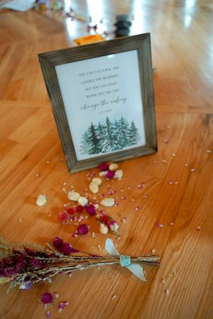A framed wedding sign with pine tree artwork sits on wooden floor with scattered flower petals.