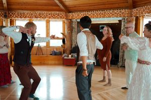 Wedding guests dance and celebrate on a wooden dance floor inside a reception hall with string lights above.