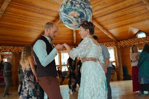 A couple dances beneath a large globe light fixture in a rustic wooden reception hall with twinkling lights.