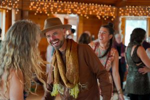 Person in brown jacket and scarf smiles while dancing in a rustic venue with string lights.