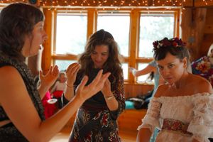 Three women in festive attire share an animated conversation in a wooden interior with natural lighting.