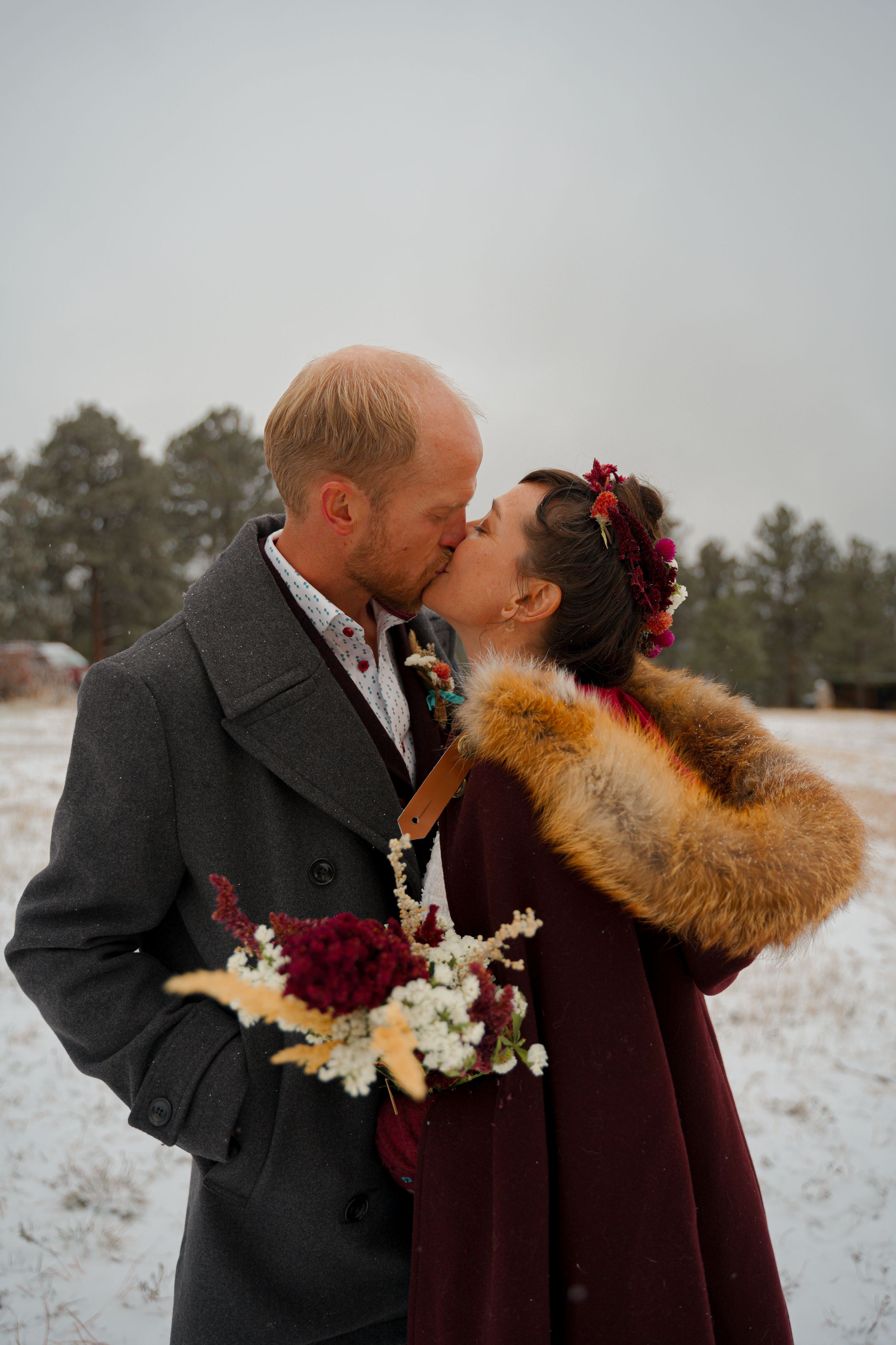 A couple shares romantic winter wedding photos in the snow with burgundy and white florals and a fur wrap.