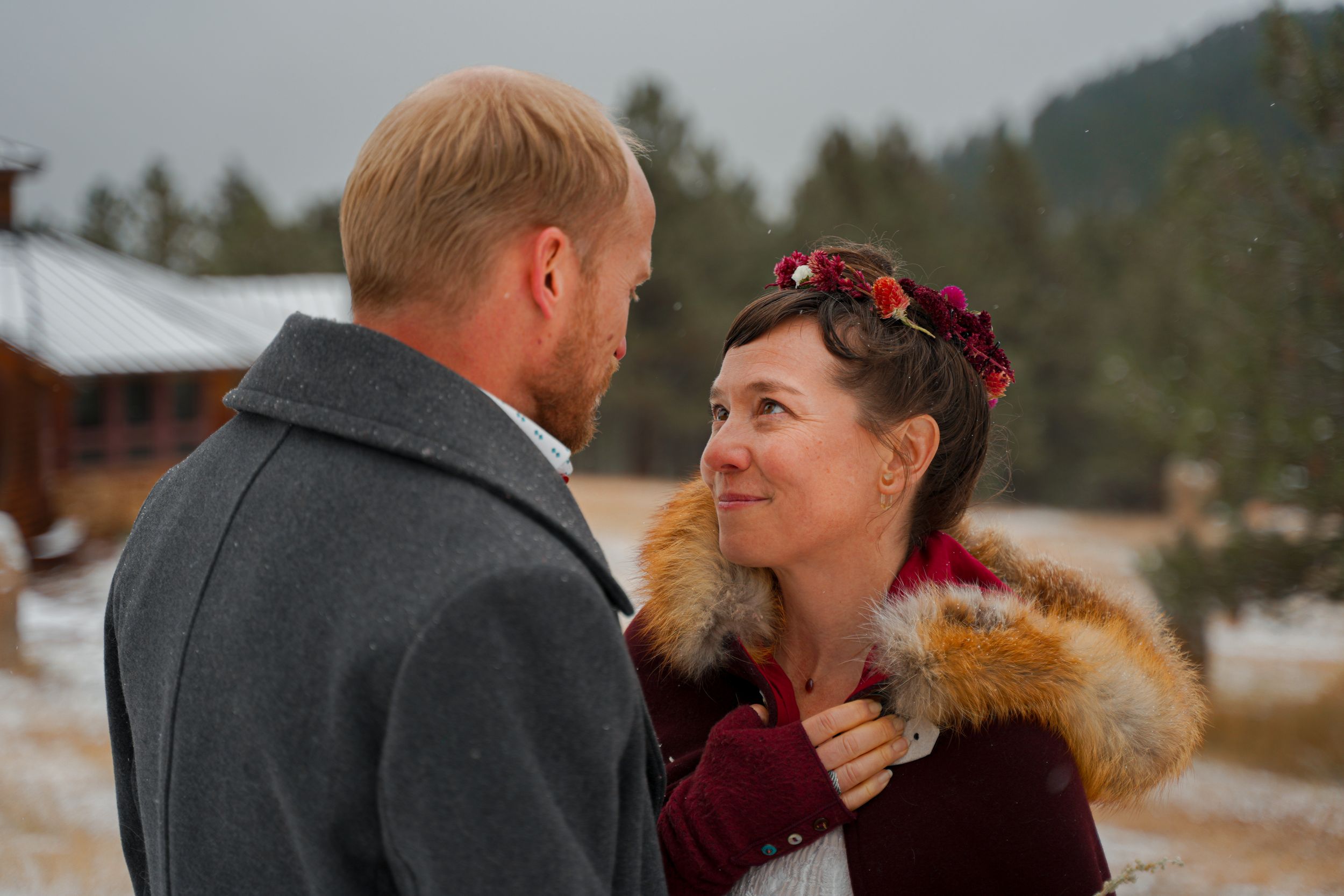 A couple shares an intimate moment outdoors in winter, wearing warm coats against a snowy mountain backdrop.