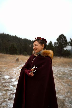 A couple in burgundy winter coats embraces in a snowy field with pine trees in the background.