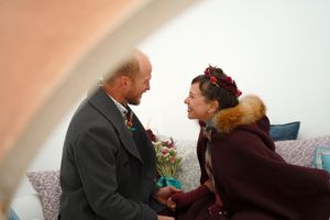 A tender exchange between two people in winter coats against a white arched backdrop.