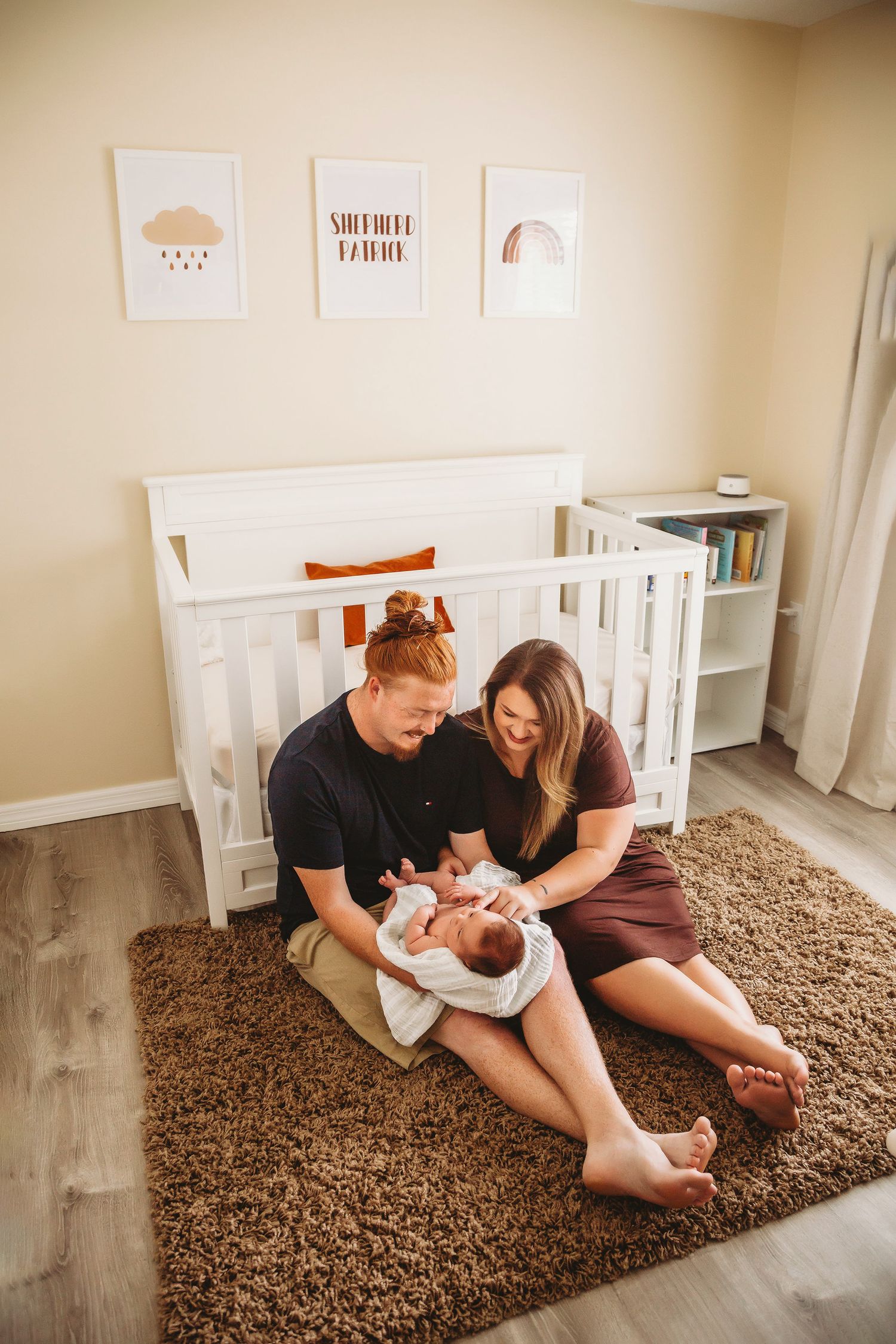 New parents with baby on brown rug in nursery with minimalist wall art.