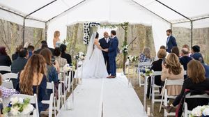 Outdoor wedding ceremony under white tent with rustic decor and guests seated on white chairs.