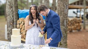 Couple cutting their wedding cake outdoors with a rustic woodpile backdrop at their forest venue celebration.