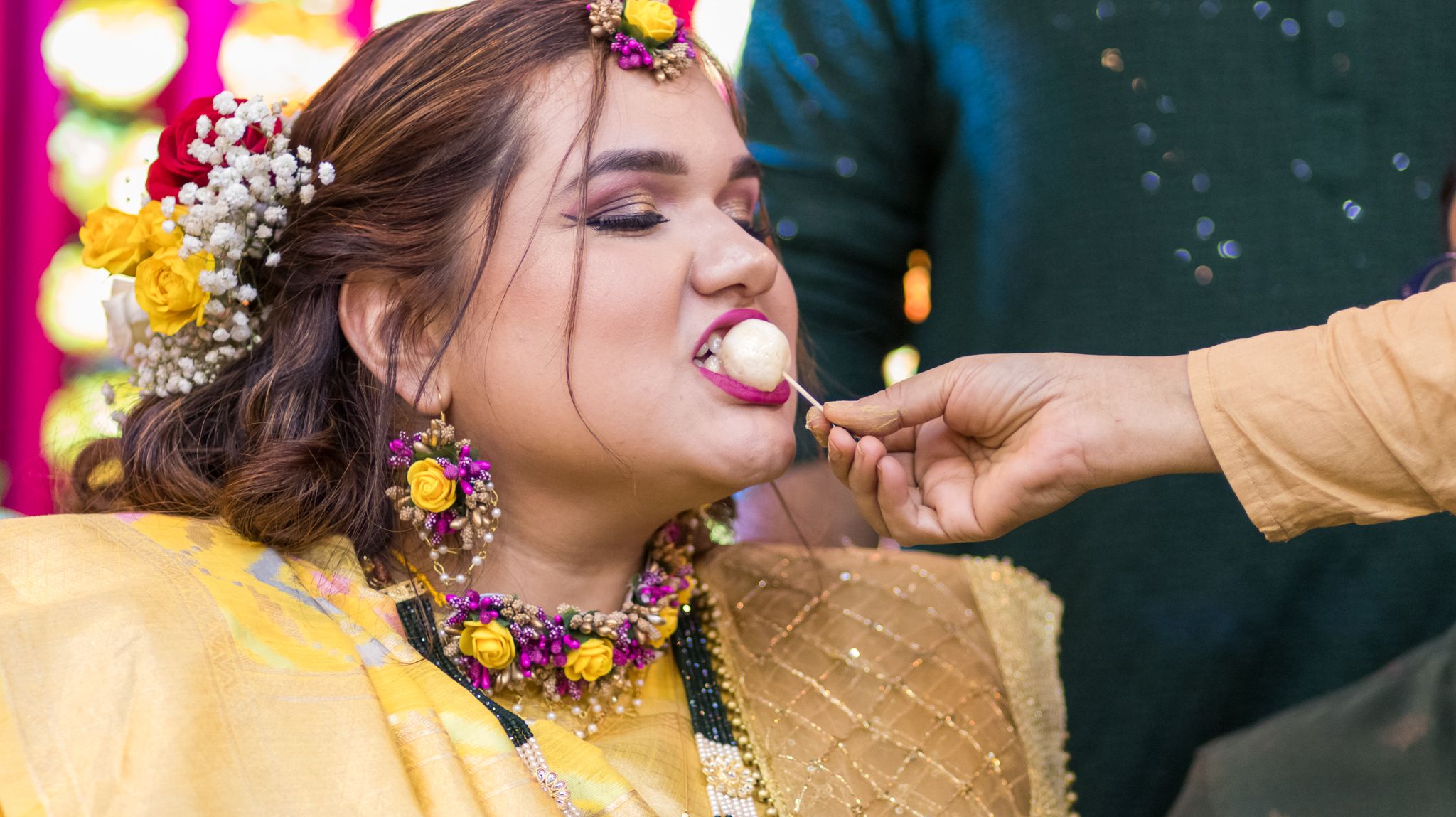 Person wearing bright yellow traditional Indian attire and floral jewelry being fed during a festive celebration.