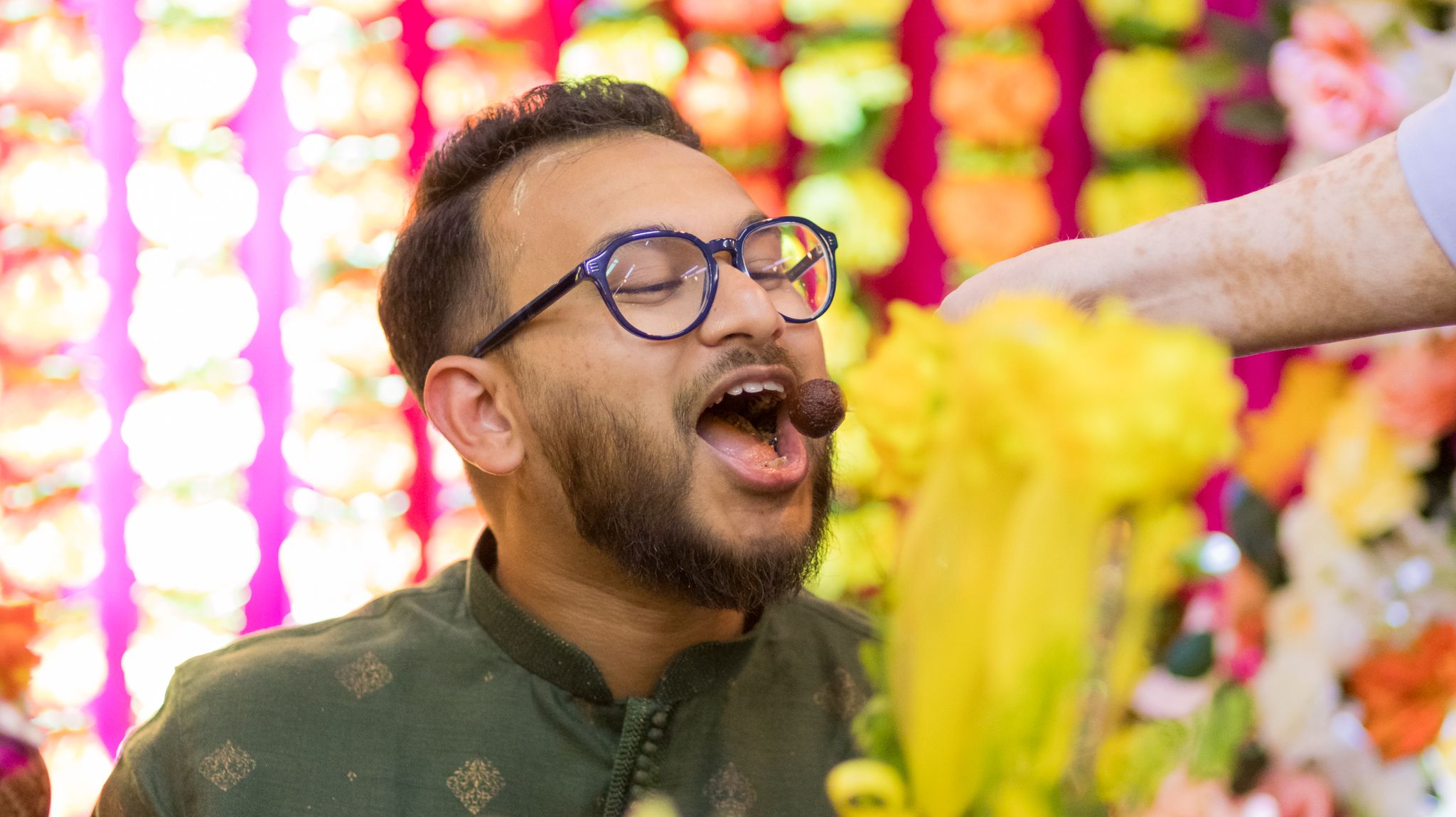 Person in glasses and traditional attire celebrating with colorful decorations in the background.