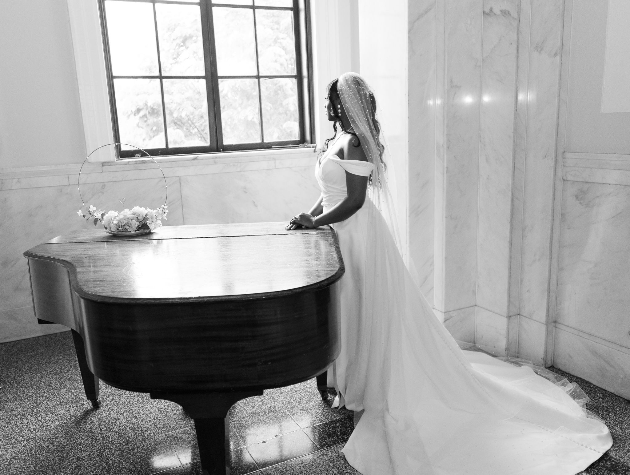 Black and white portrait of bride posing beside grand piano in elegant wedding gown.