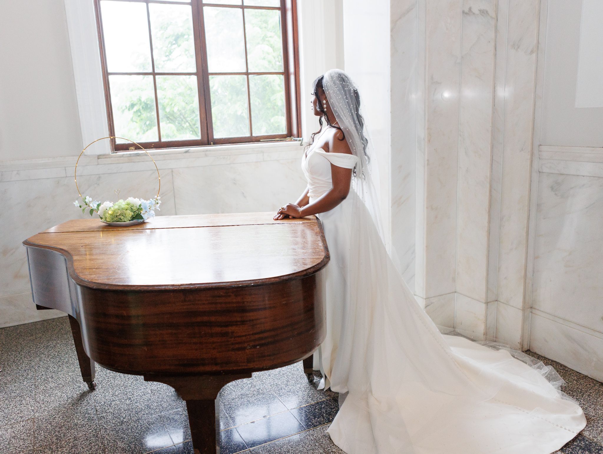 Side view of bride in white gown standing beside vintage piano in bright room.