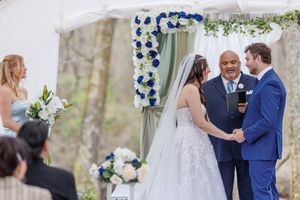 Beautiful outdoor wedding ceremony with blue floral arch decorations and guests seated in nature.