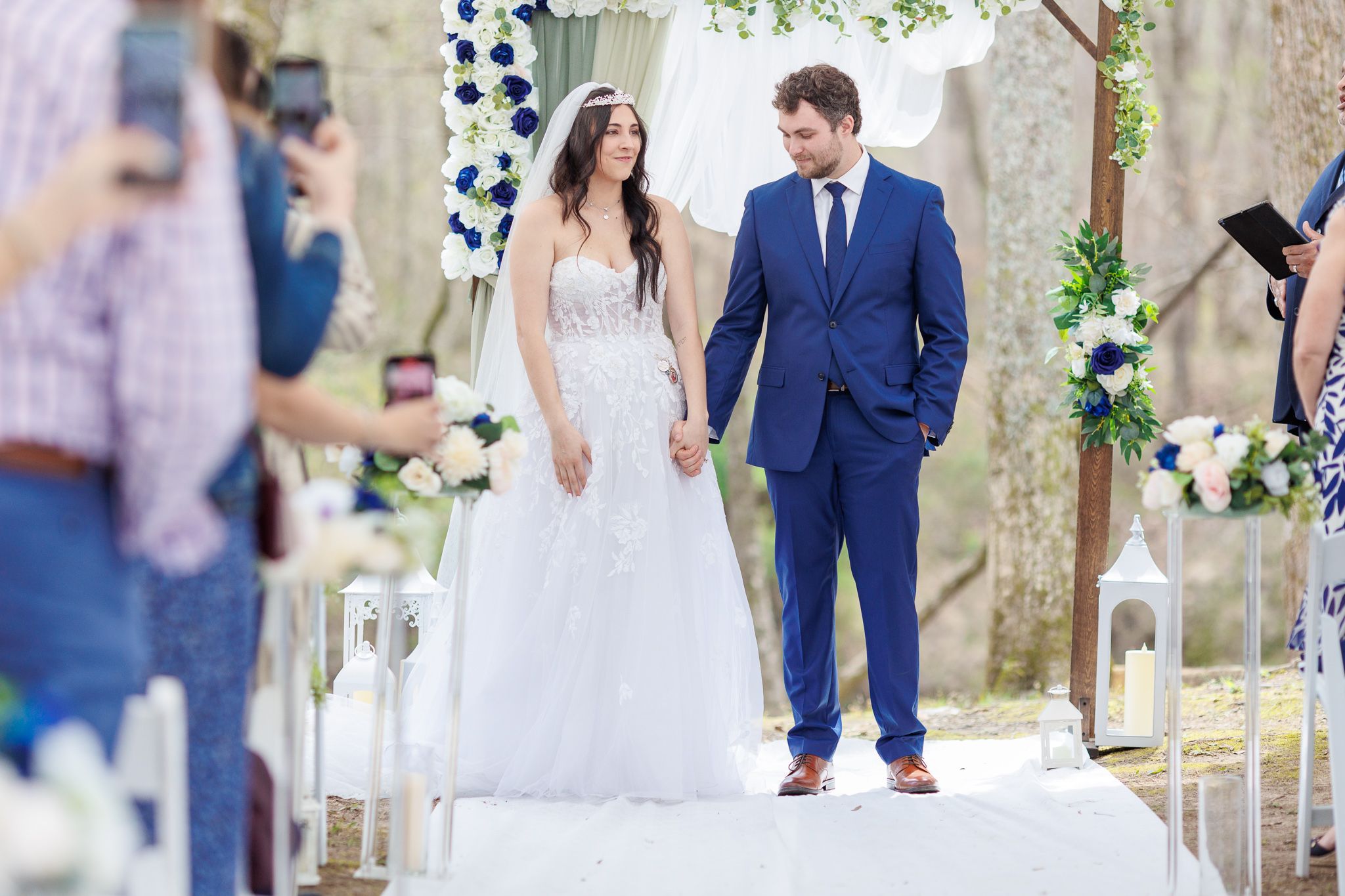Wedding couple walking down white aisle runner during outdoor forest ceremony with guests watching.