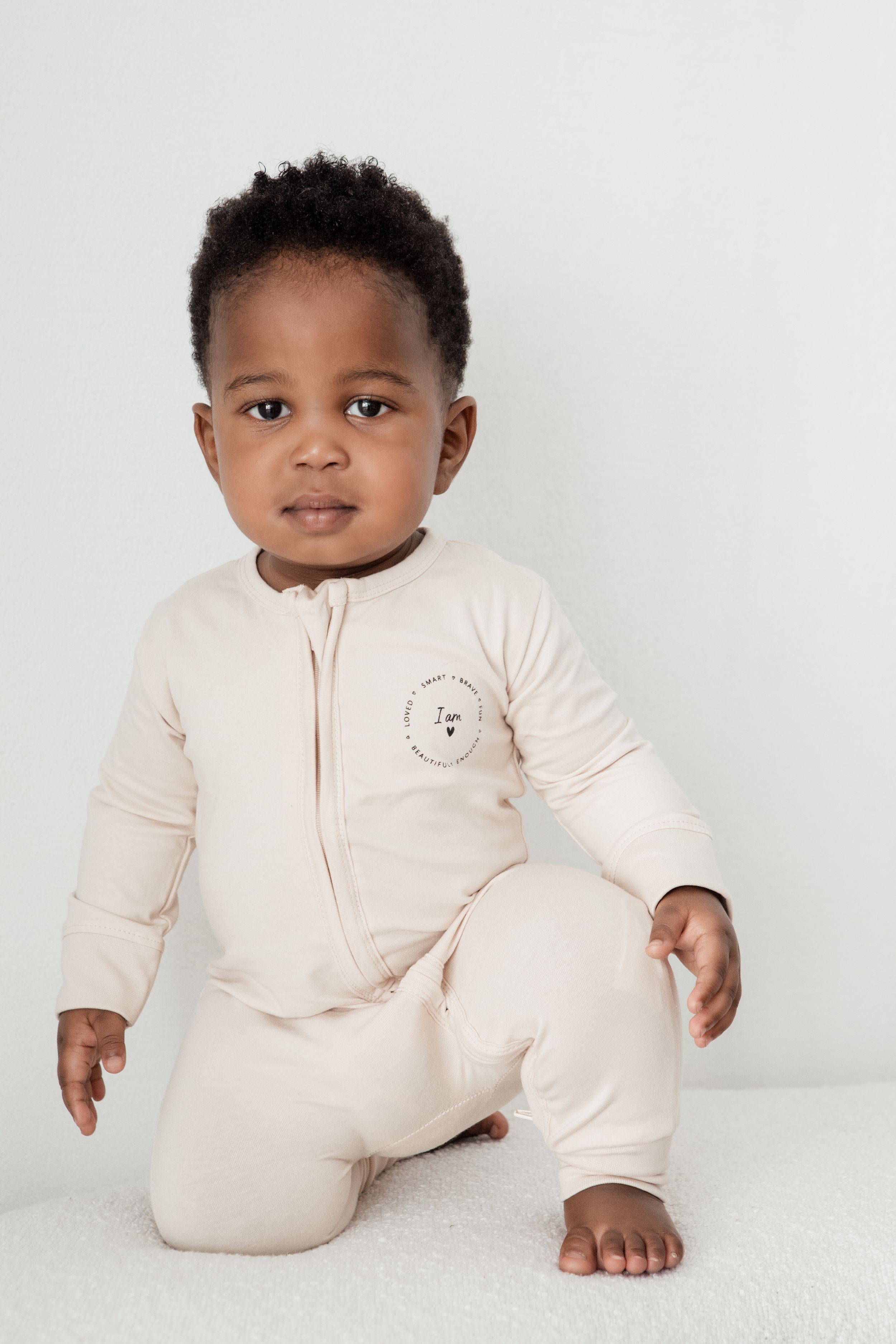 Toddler wearing cream-colored zip-up onesie sitting on white background.