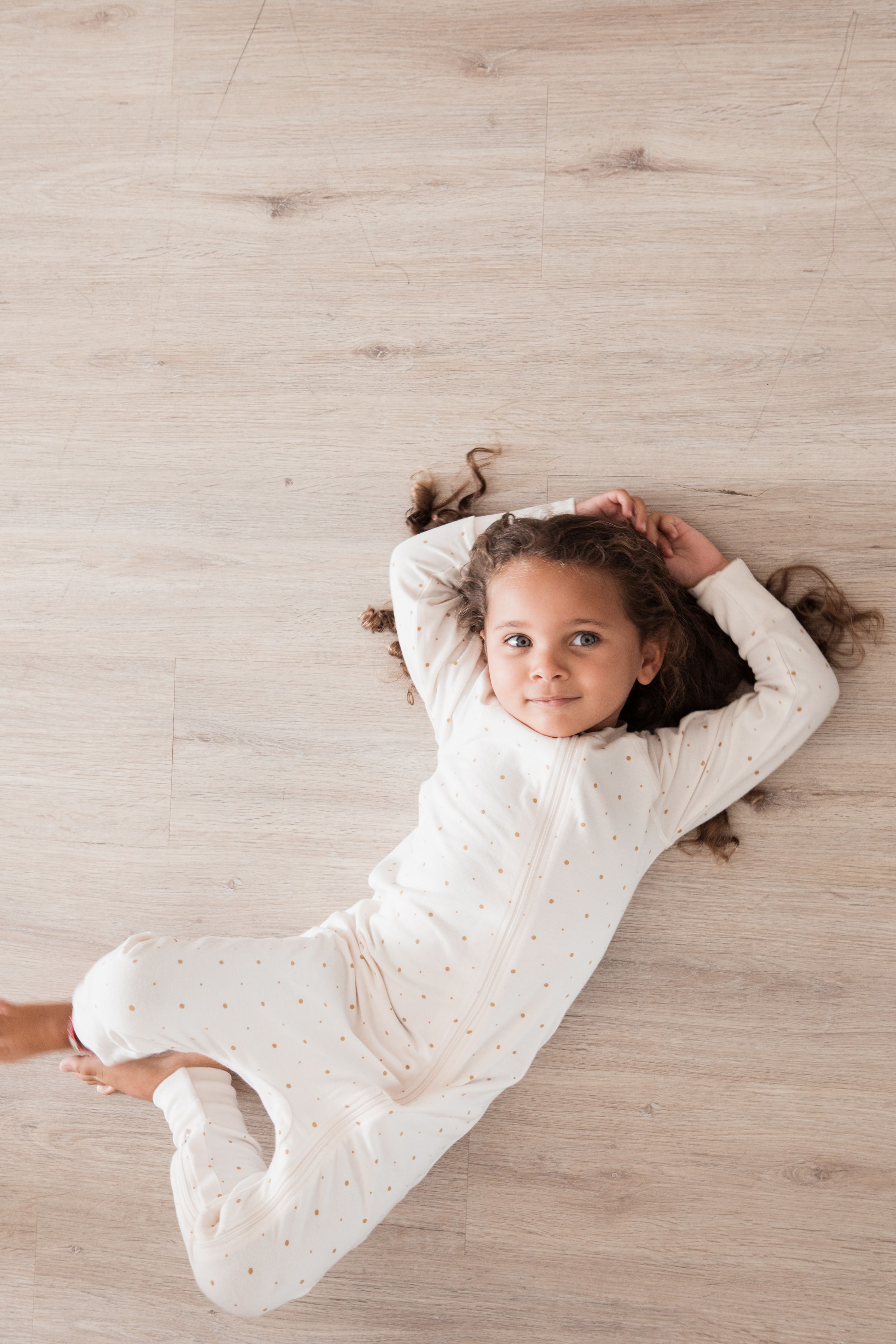 Child in white dotted pajamas lying on light wooden floor.