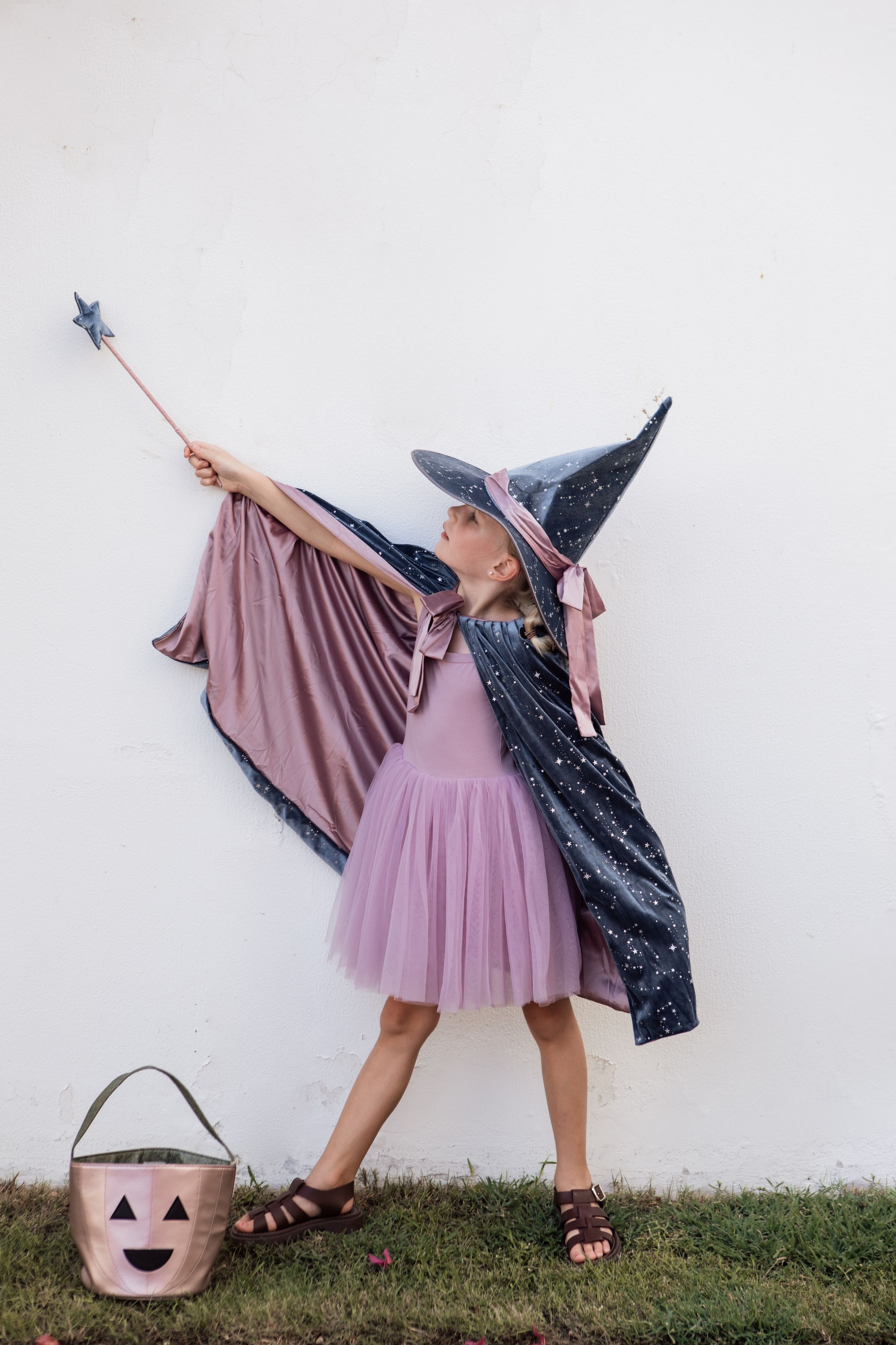 Child in pink tulle dress and witch costume posing with wand against white wall.