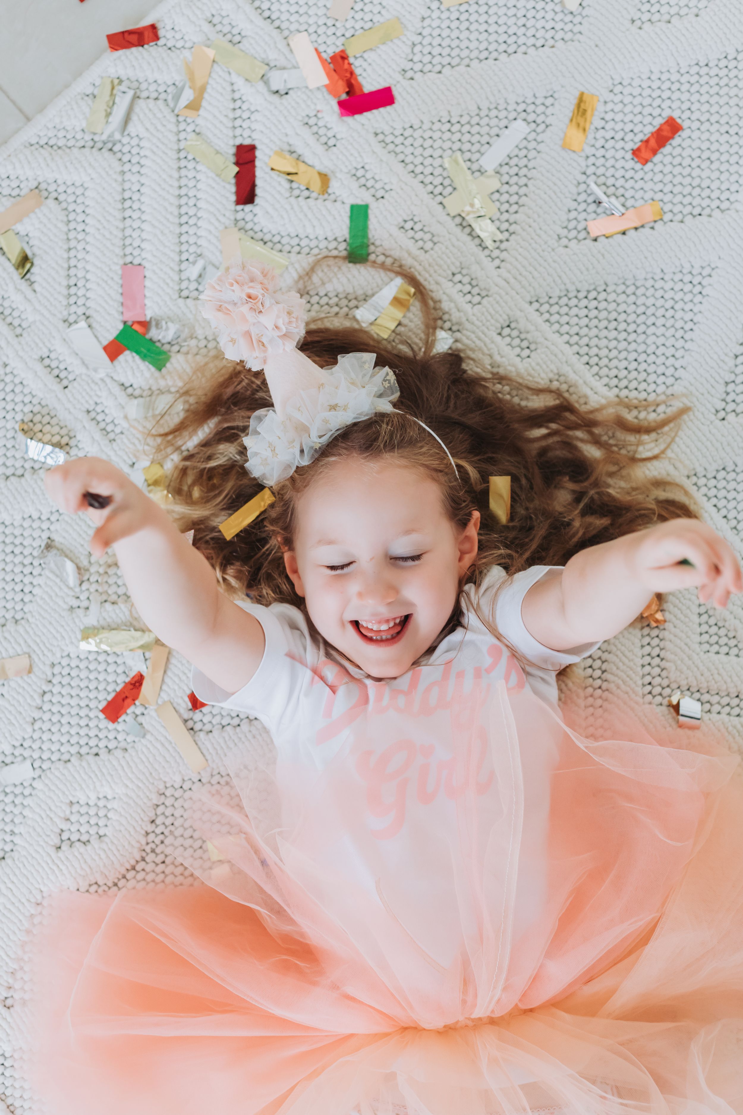Child in pink tutu dress laying on ground surrounded by confetti in joyful celebration pose.