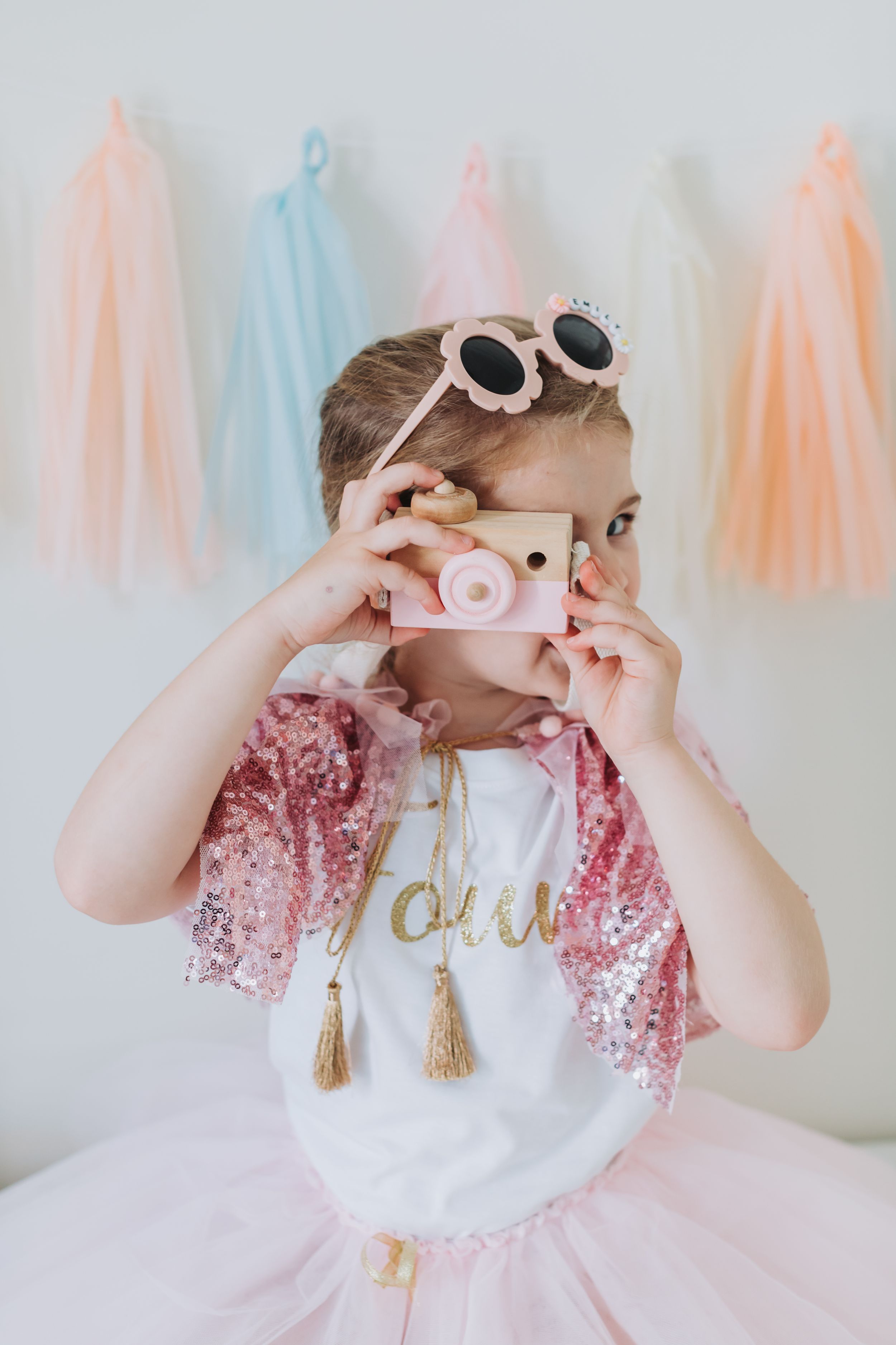 Child wearing pink tutu and sunglasses while holding toy camera against pastel backdrop.