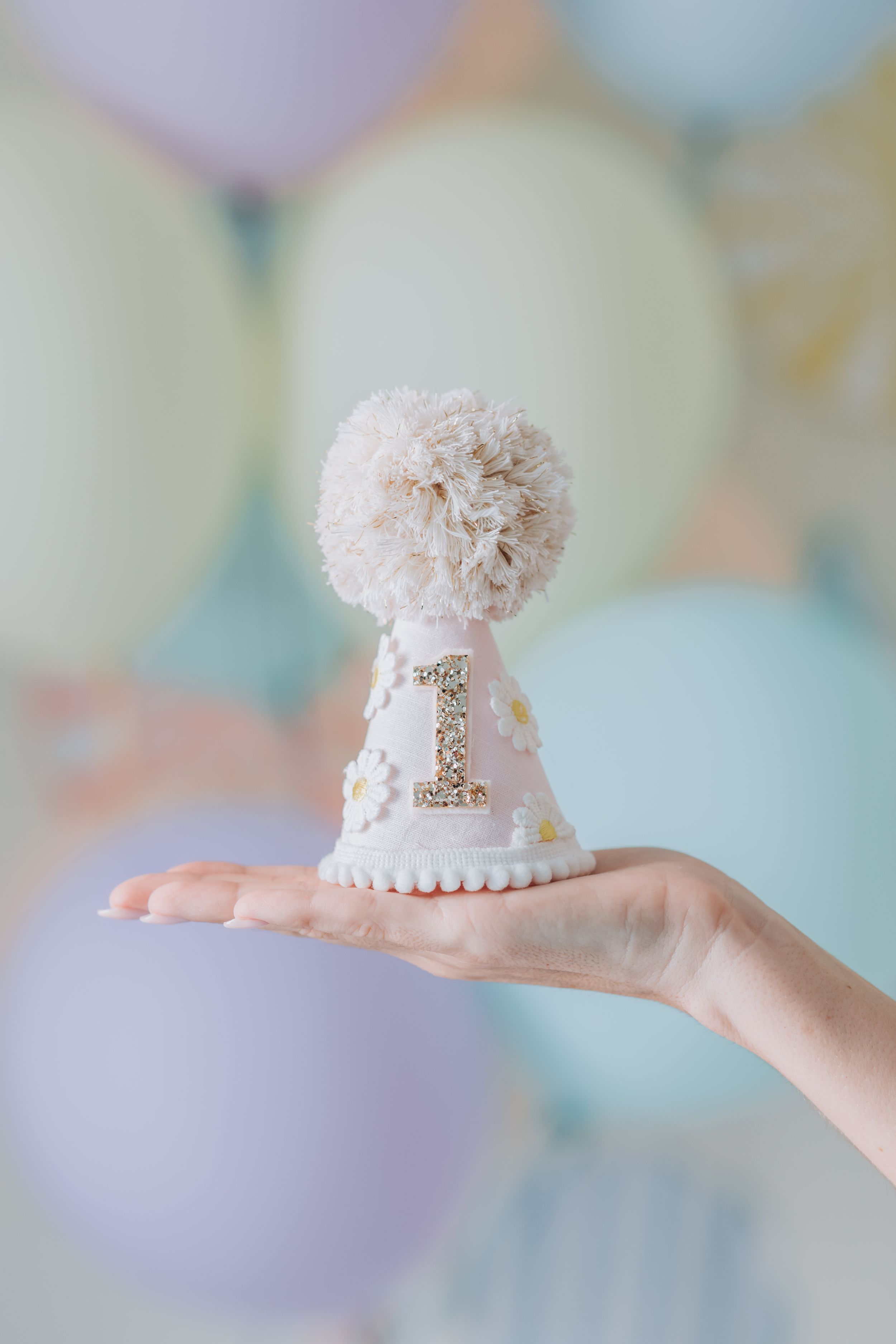 White decorative party hat with number 1 and pom pom being held against pastel balloon background.