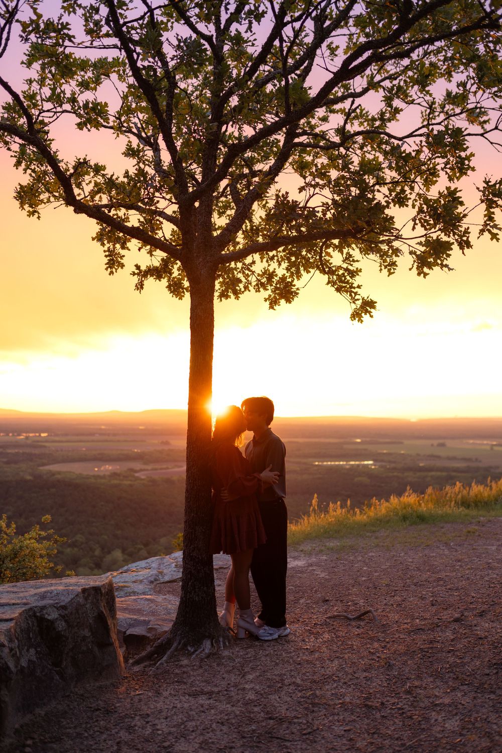 Couple sitting under a large tree silhouetted against dramatic sunset landscape.