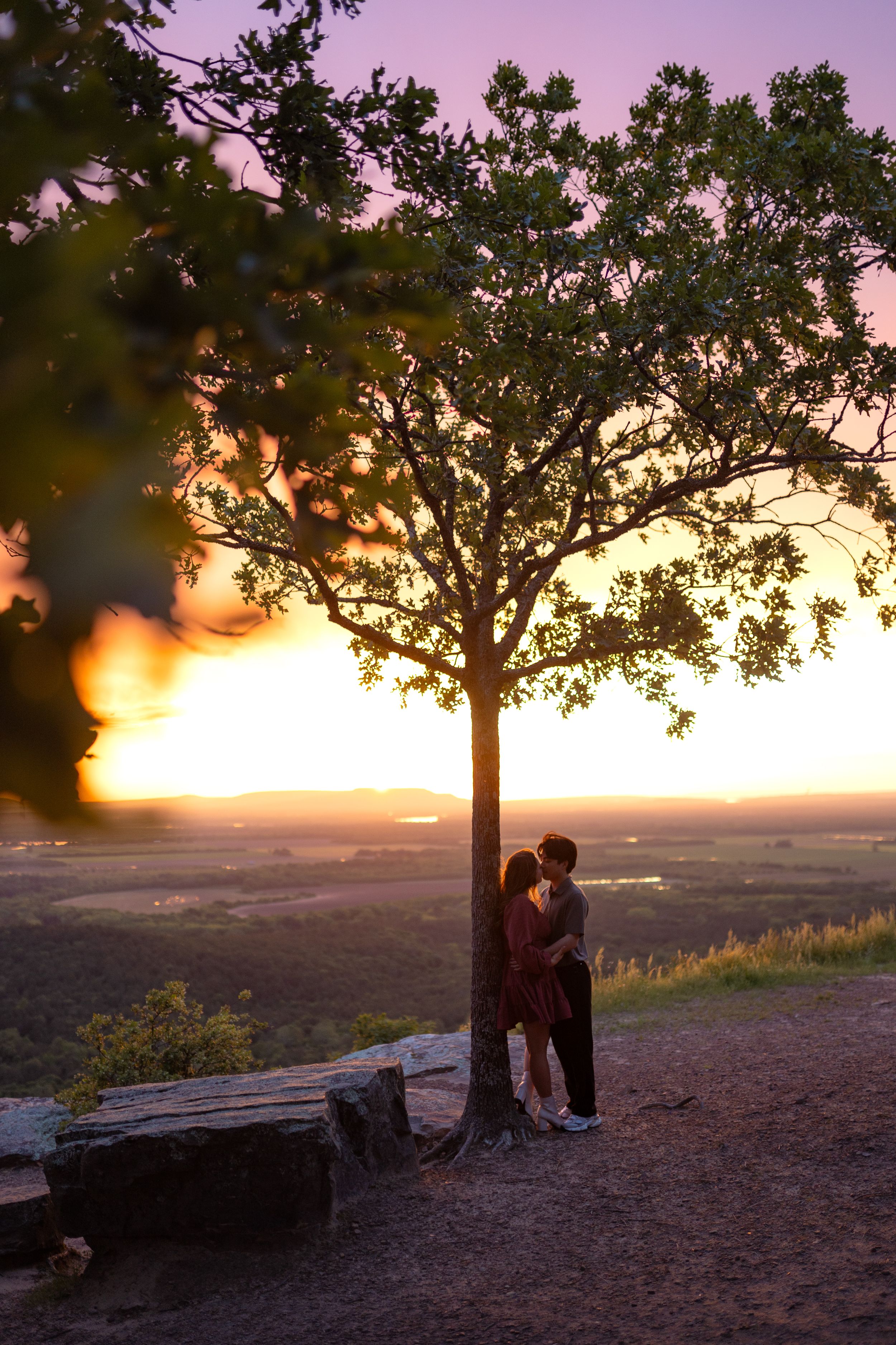 Two people nestled under a tree overlooking a vast landscape at sunset.