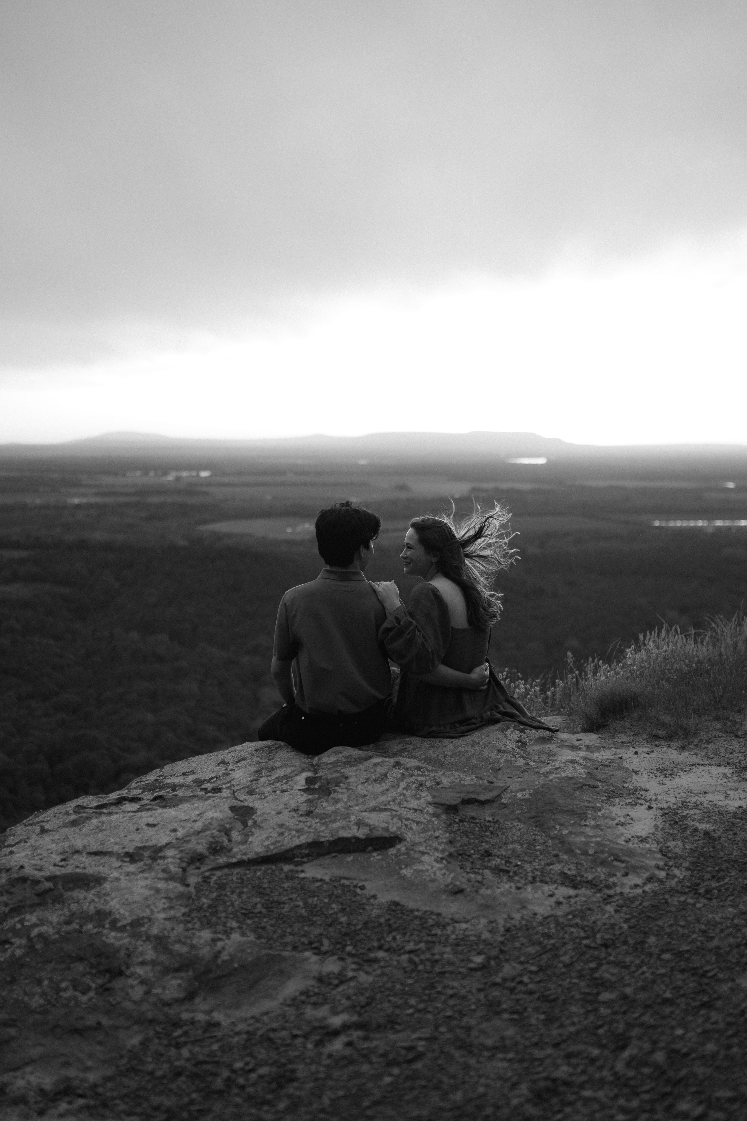 Black and white photo of couple lying on cliff edge overlooking scenic vista.