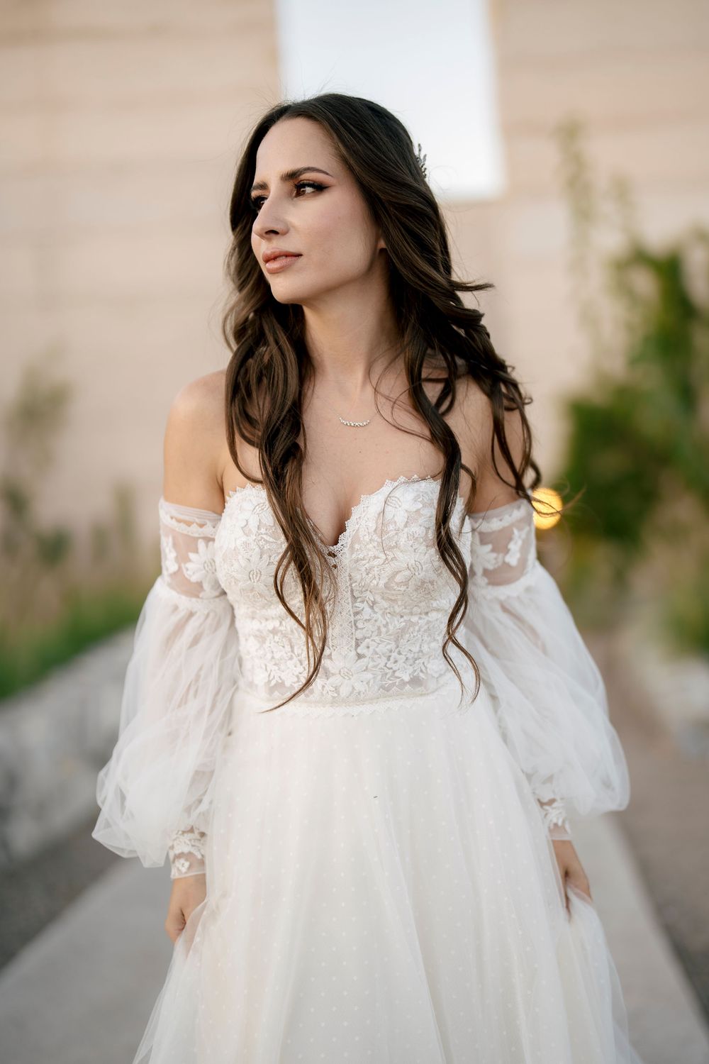 A bride in an off-shoulder white lace wedding dress poses outdoors with windswept hair.