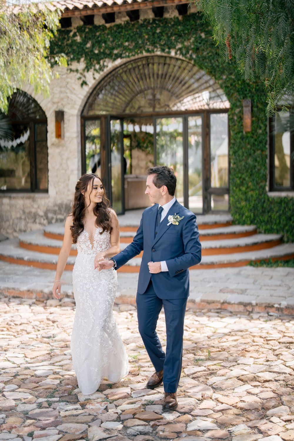 A couple walks together on a cobblestone path in front of an elegant ivy-covered Mediterranean-style building.