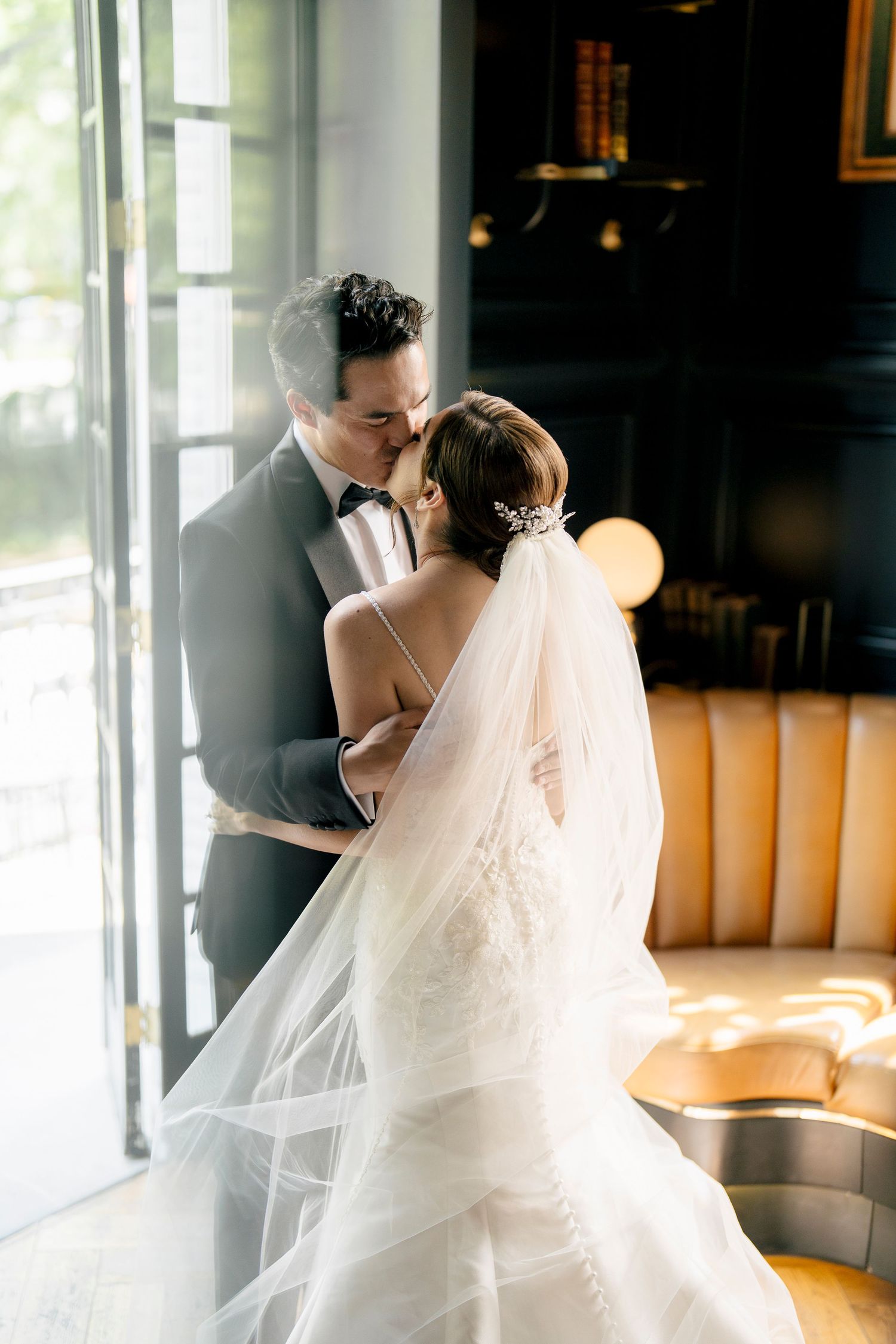 Sunlit wedding couple sharing tender moment near large windows.