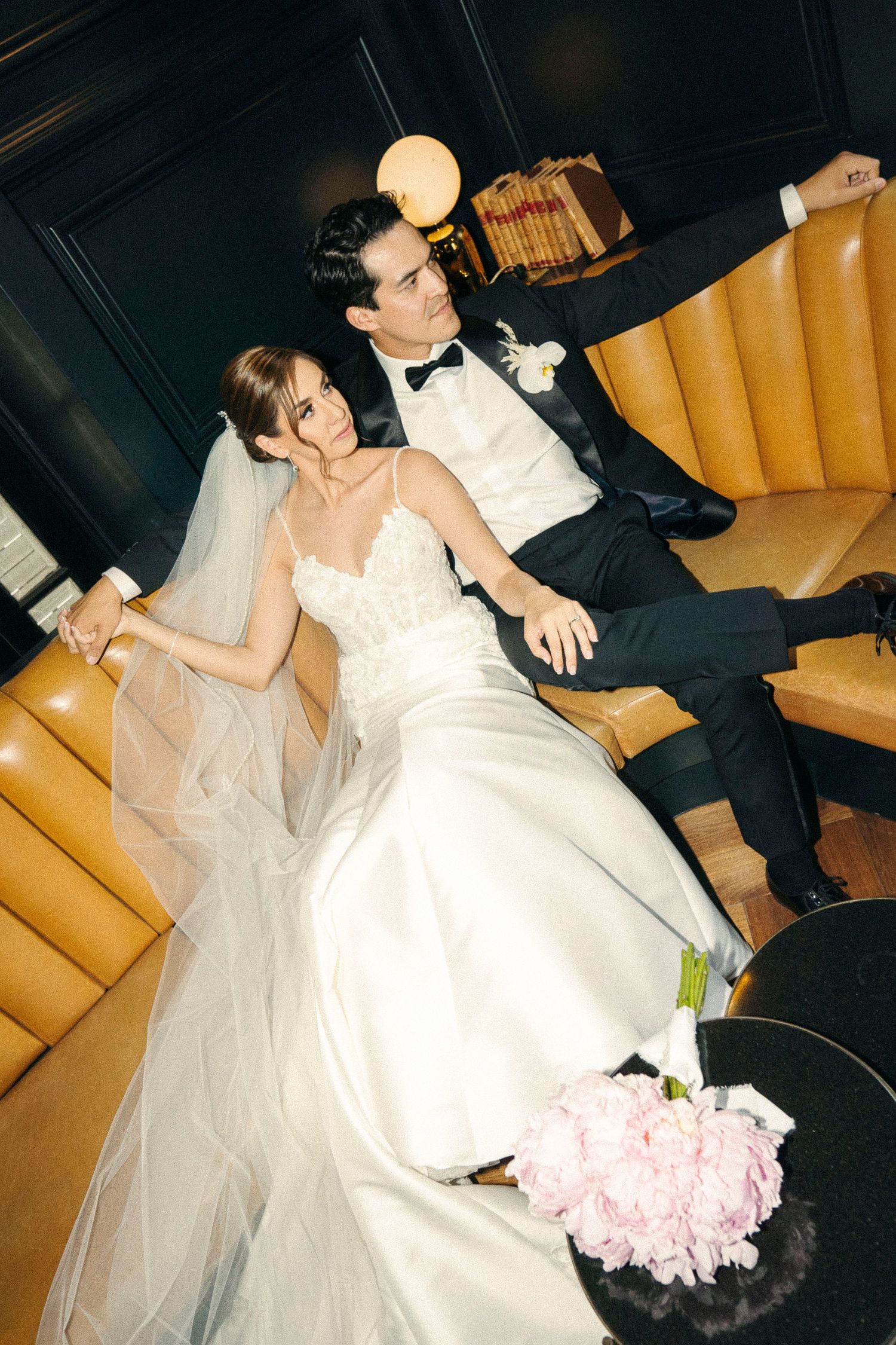 Newlyweds relaxing on tan leather couch with pink peony bouquet.