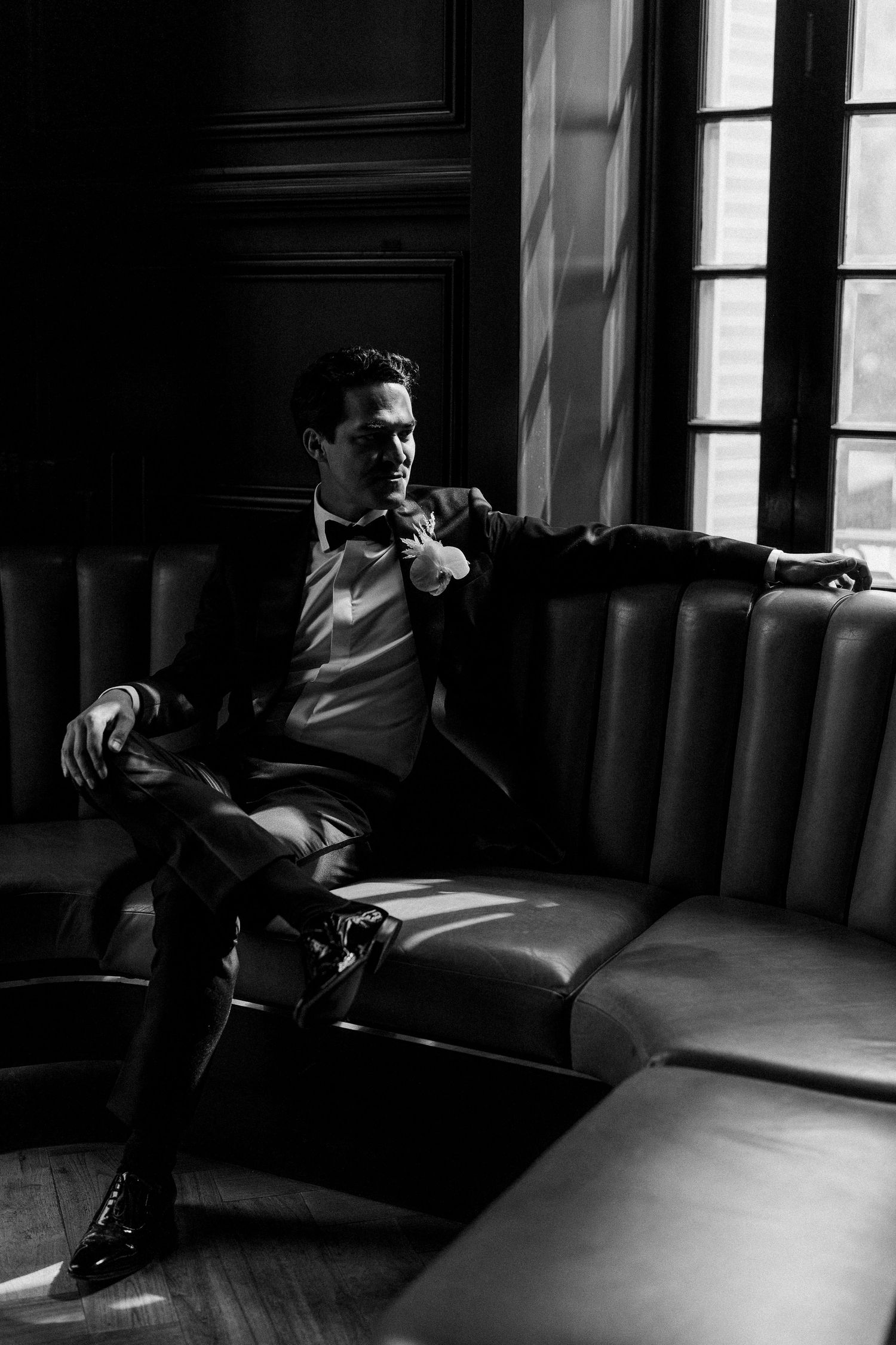 Dramatic black and white portrait of groom lounging on vintage leather seating.