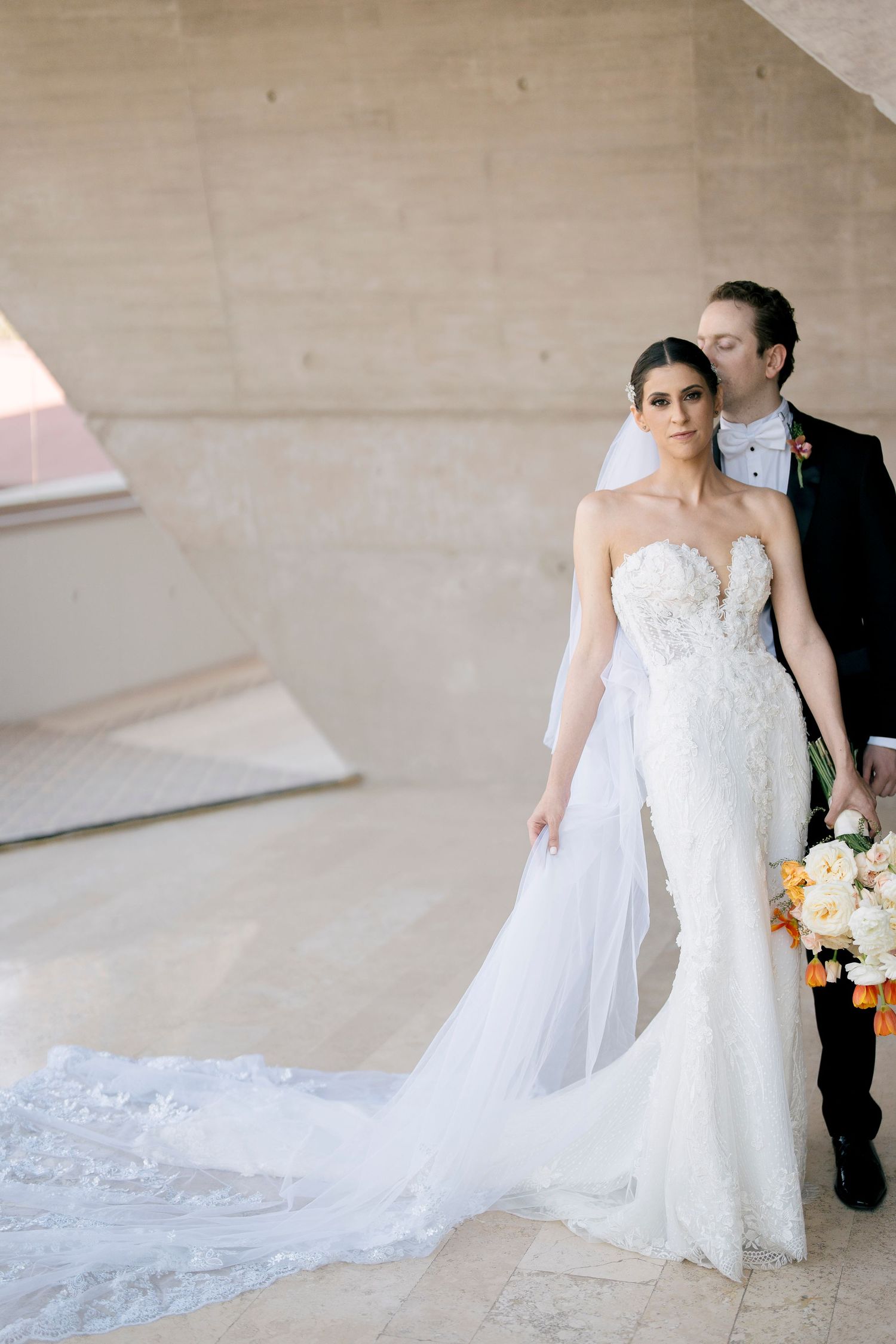Bride in flowing white lace gown with dramatic train against neutral wall.