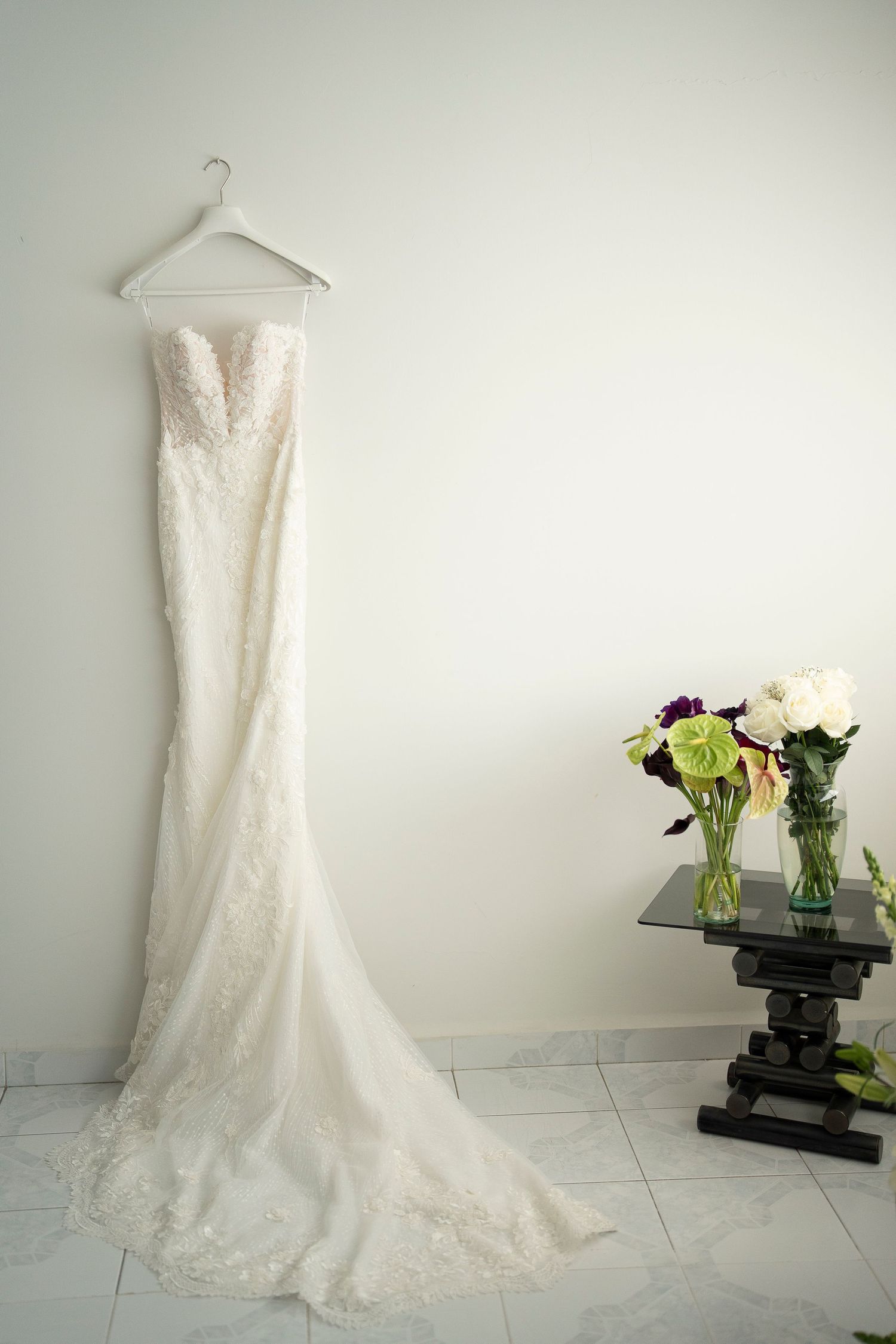 Wedding dress hanging display with delicate lace details and floral arrangements against white wall.