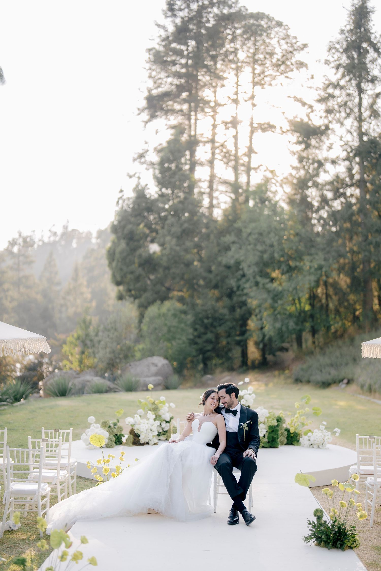 A serene outdoor wedding venue setup with white chairs on a hillside surrounded by tall evergreen trees.
