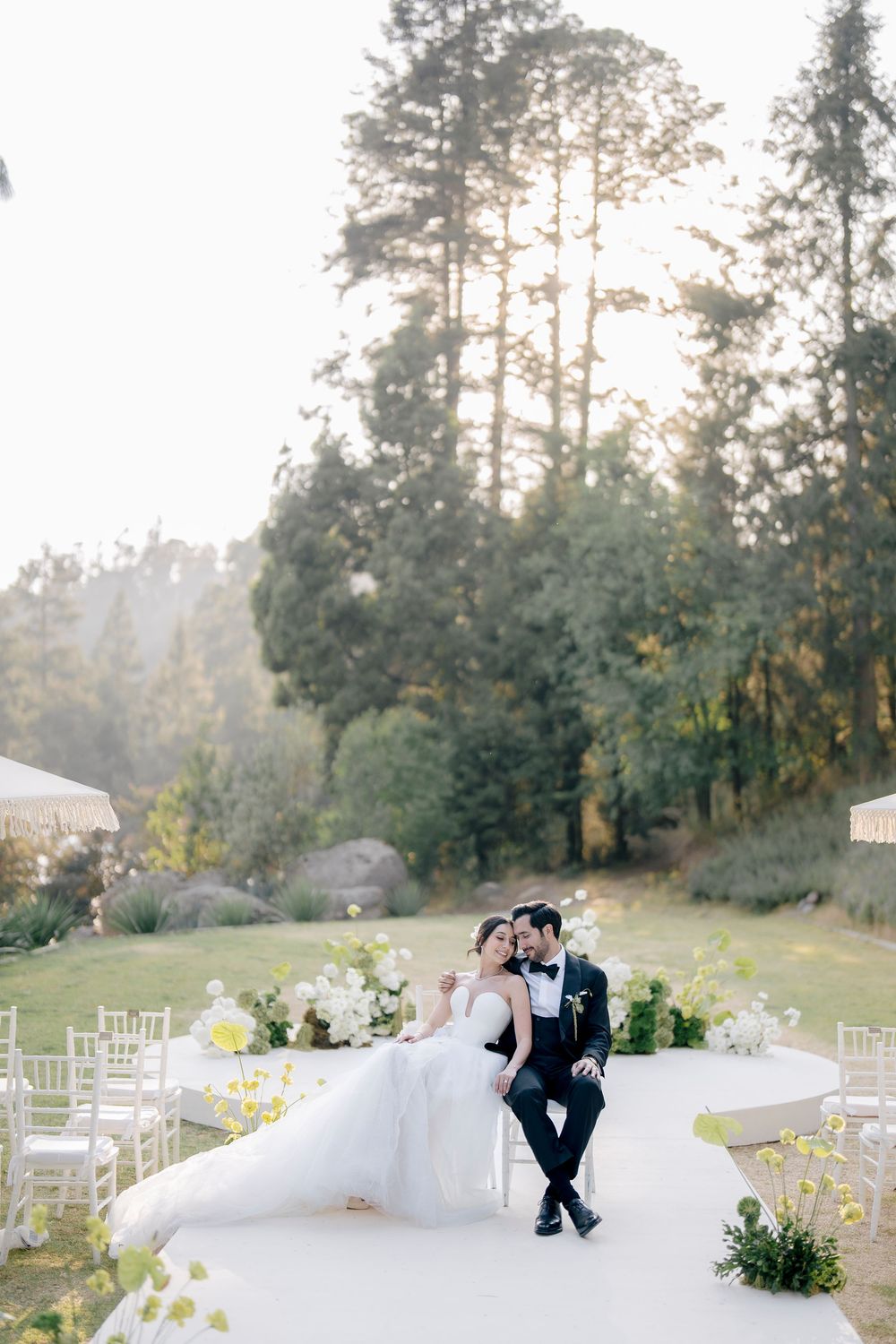 A serene outdoor wedding venue setup with white chairs on a hillside surrounded by tall evergreen trees.