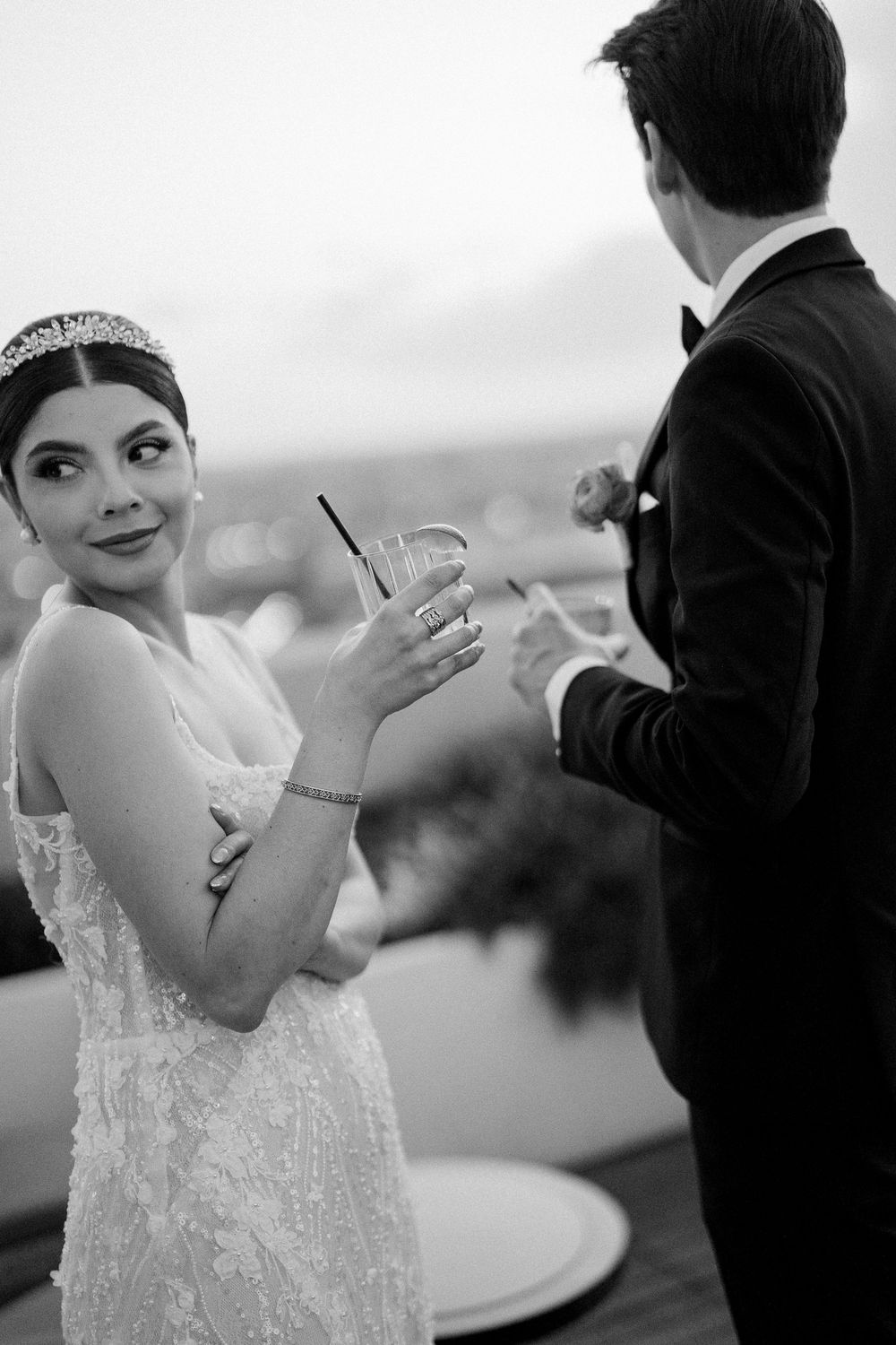 Black and white photo showing a bride in a lace dress holding a cocktail while celebrating with her groom outdoors.