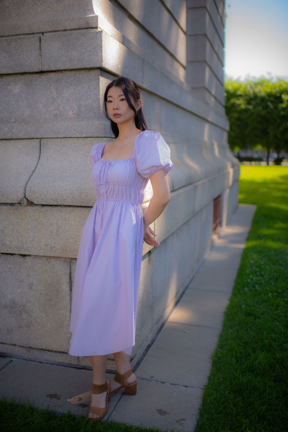 Full-length portrait in a lilac dress with puff sleeves against a stone building exterior.