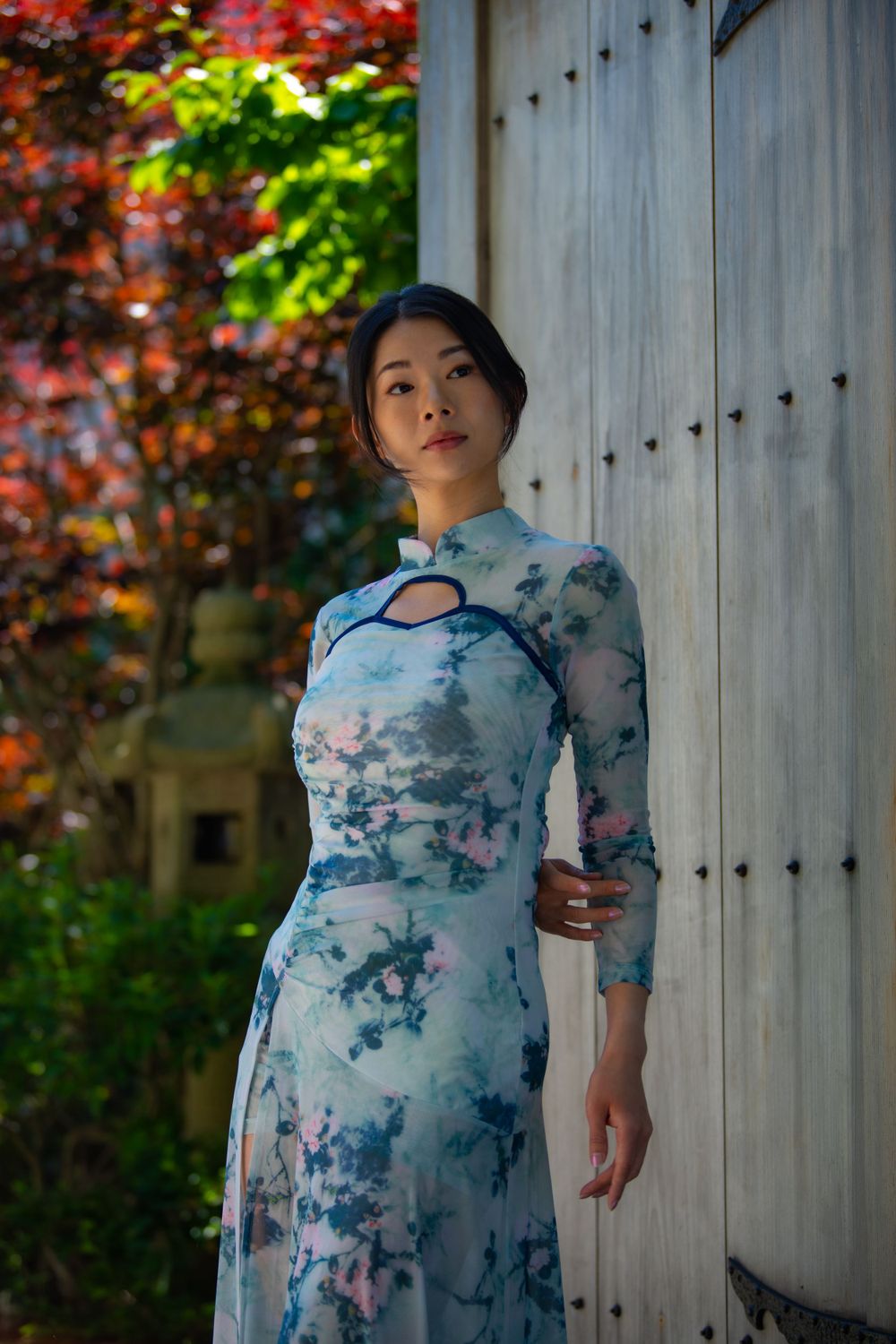 An elegant pose in a blue floral qipao-style dress against a wooden wall with red maple leaves.