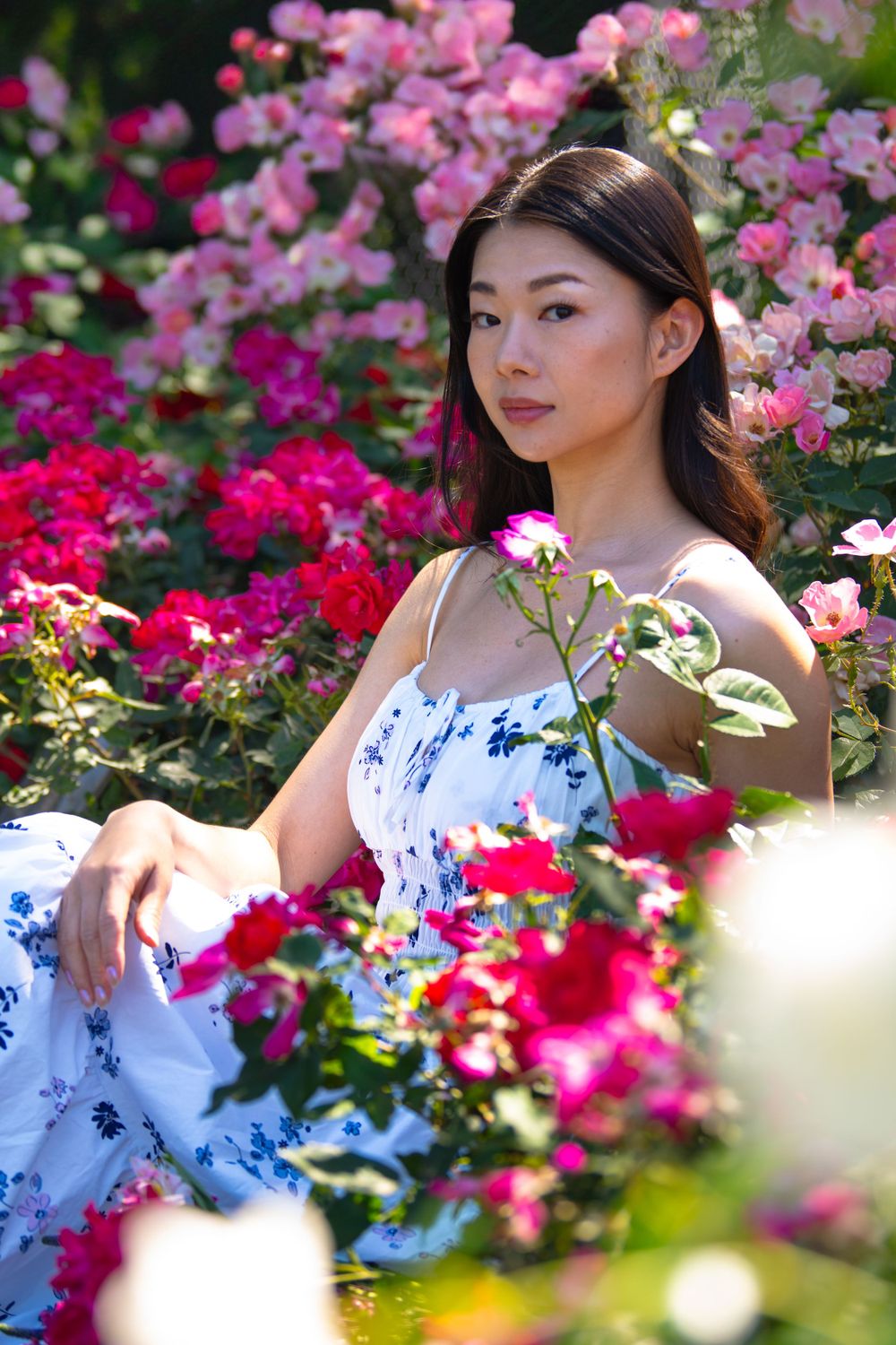 Model in blue and white dress surrounded by vibrant pink and red flowers in garden setting.