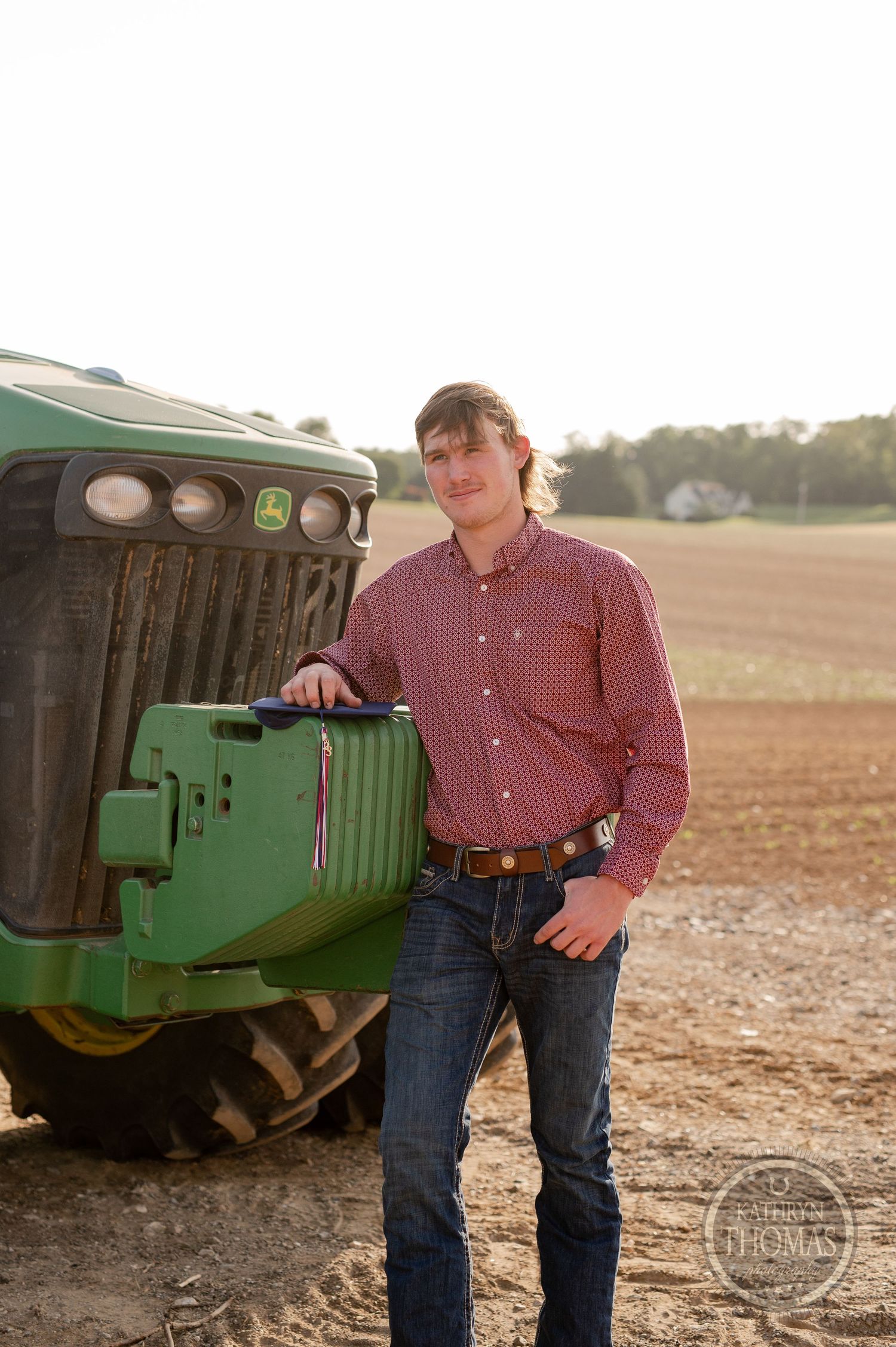 Person in checkered shirt poses beside green John Deere tractor in field.